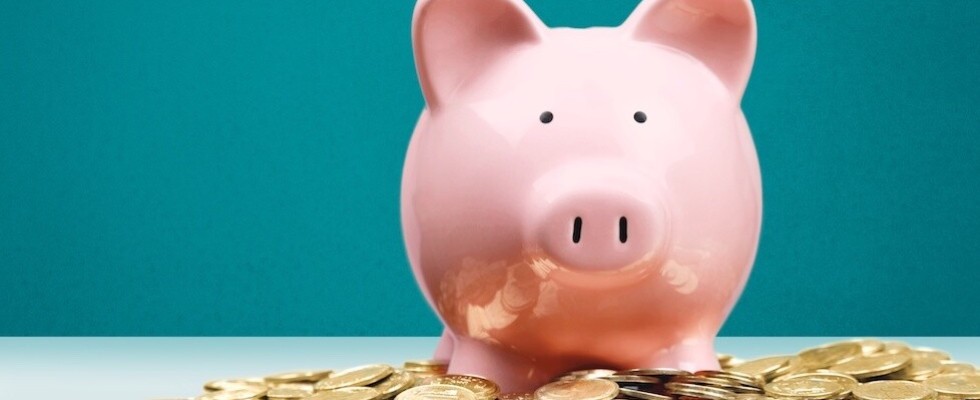 A pink piggy bank on a table in front of a blue wall with gold coins surrounding it.