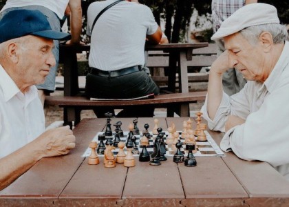 Two elderly men in white shirts playing a game of chess outside on a wooden table.