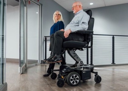 An image of a man sitting in a power seat elevation wheelchair with a woman walking beside him.  