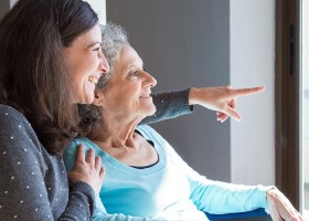 An older woman is being cared for by her middle-aged daughter and they are looking out the window.