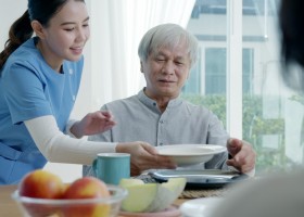 A young woman caregiver is bringing a tray of breakfast food to an older gentleman in his home.