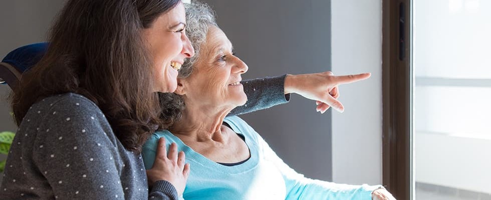 An older woman is being cared for by her middle-aged daughter and they are looking out the window.