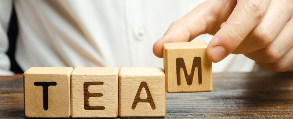 A man sits behind a wooden table with three wooden blocks in front of him with the letters T, E, and A on them. He is setting down another with the Letter M, to spell out the word "Team".