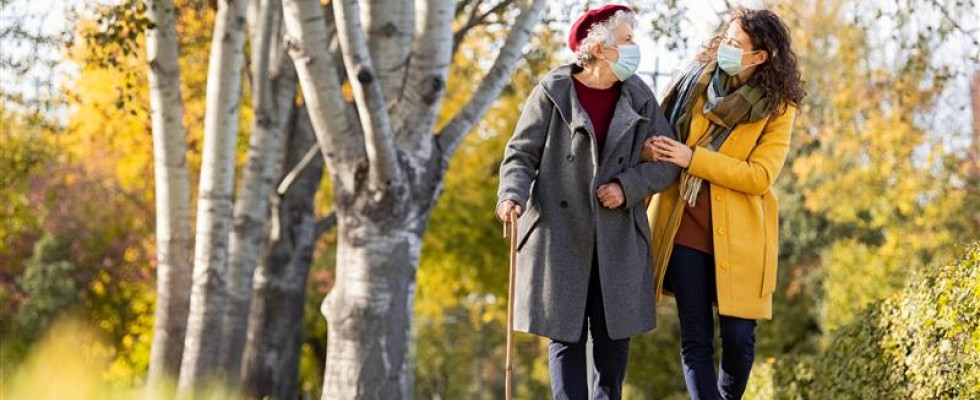 An elderly woman walks with a cane in one hand and her other arm linked around her caregiver's arm