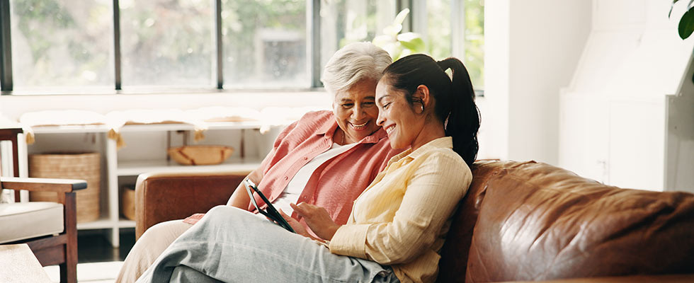 An elderly woman and her daughter sit on a couch smiling while looking at an iPad.