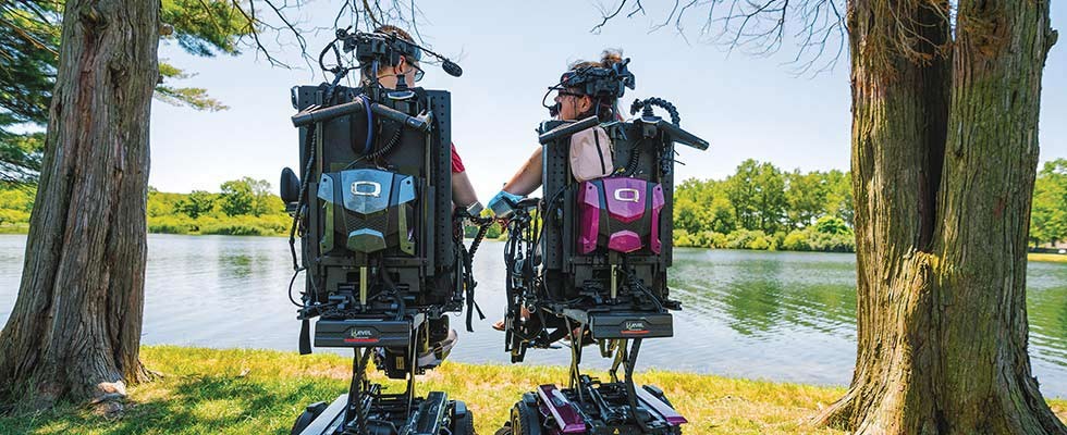 Two people sitting in medical assist chairs by a sunny lake
