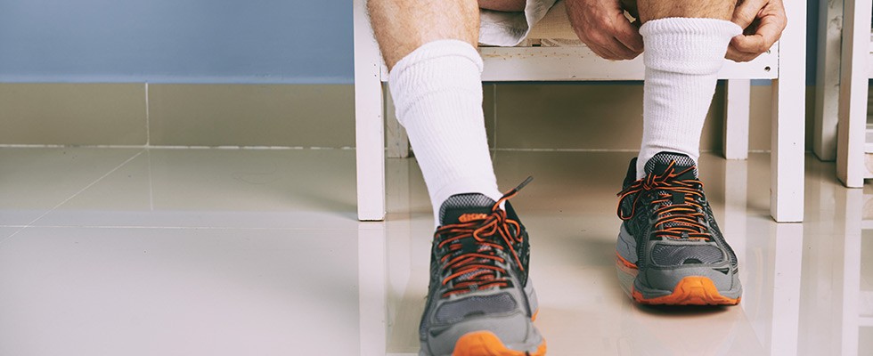 A man leans over to adjust his socks. He is sitting in a white chair while wearing running shoes