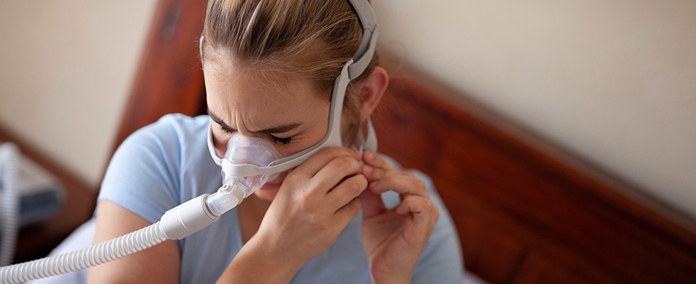 A woman puts on a CPAP machine and appears to be frustrated or in pain.