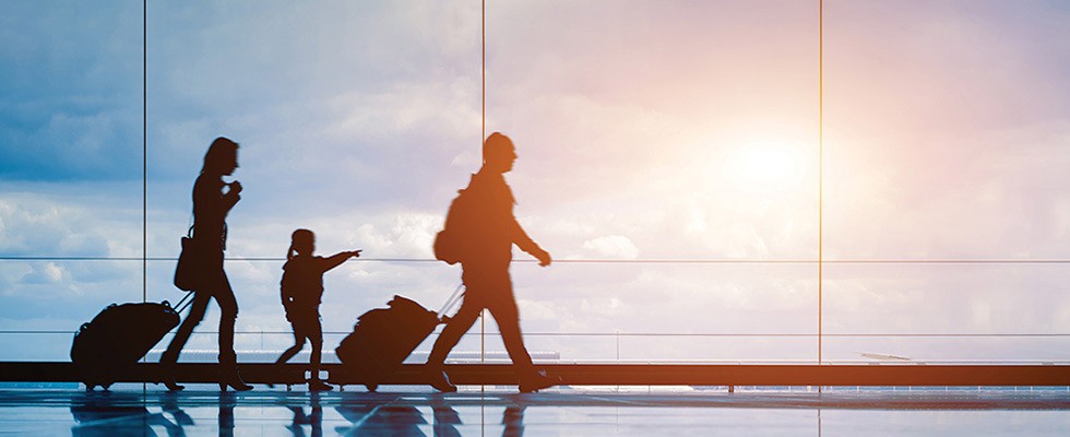 A family of three walking through an airport with their luggage as the sunrise shines in the background.