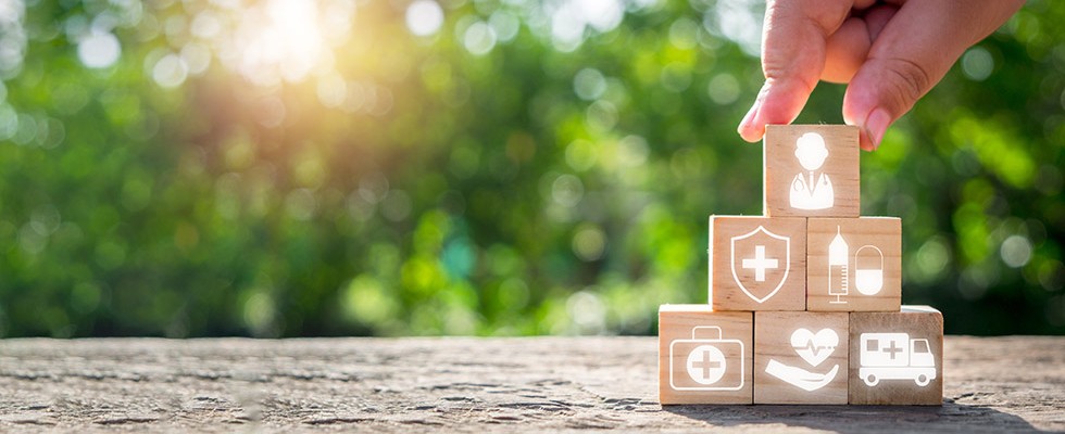 In the foreground a hand is stacking six blocks into a pyramid. On each block are different symbols of health care, including: a doctor, an ambulance, a syringe and pill, a medical shield, a first aid kid and a hand with a heart and pulse over it. In the background are blurry trees.