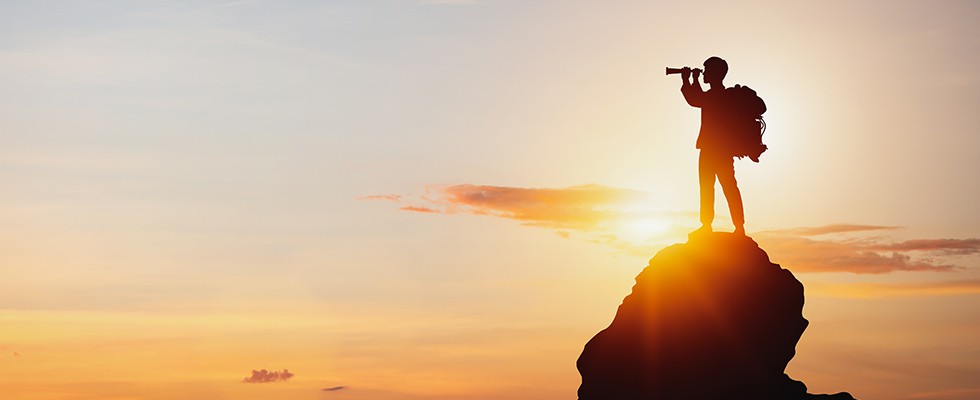A man standing on a rock with a telescope looking out into sunset.