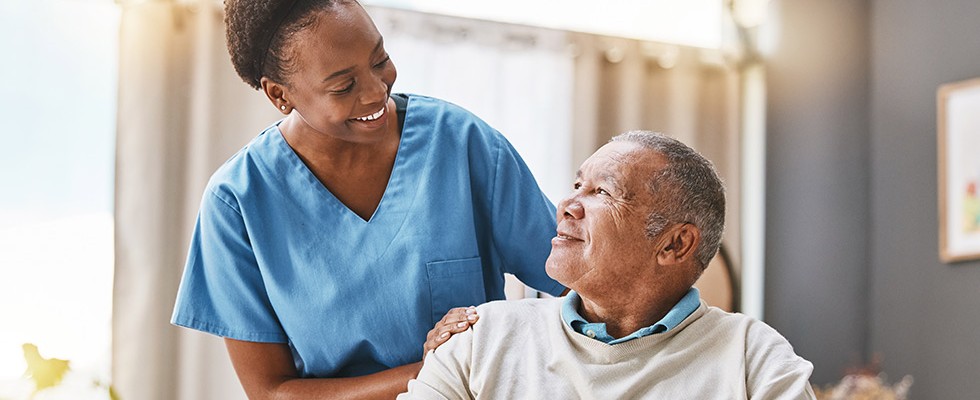 A caregiver smiles at a patient sitting down in a room.