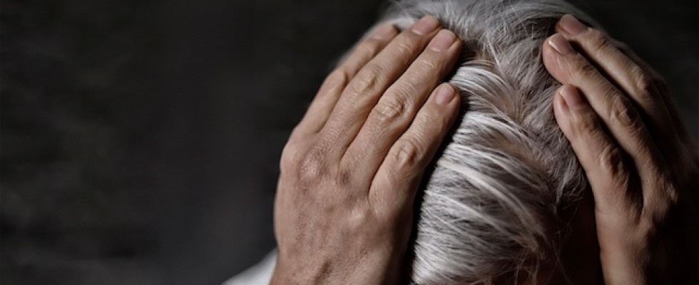 An elderly person with grey hair holds onto the top of their head.