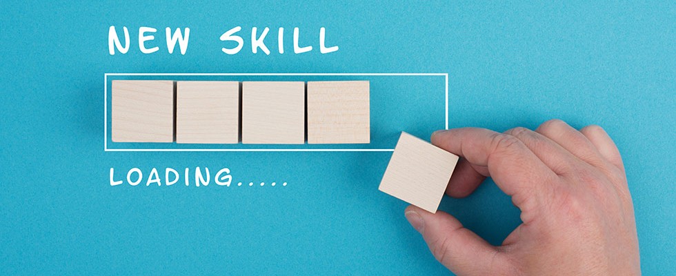 The words "New Skill" are written over an outline of a box with four wood blocks in it resembling a loading bar. Below the box are the words "Loading..." A hand is in the midst of placing another block into the box, resembling the loading being close to complete.
