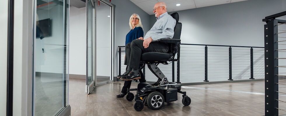 An image of a man sitting in a power seat elevation wheelchair with a woman walking beside him.  