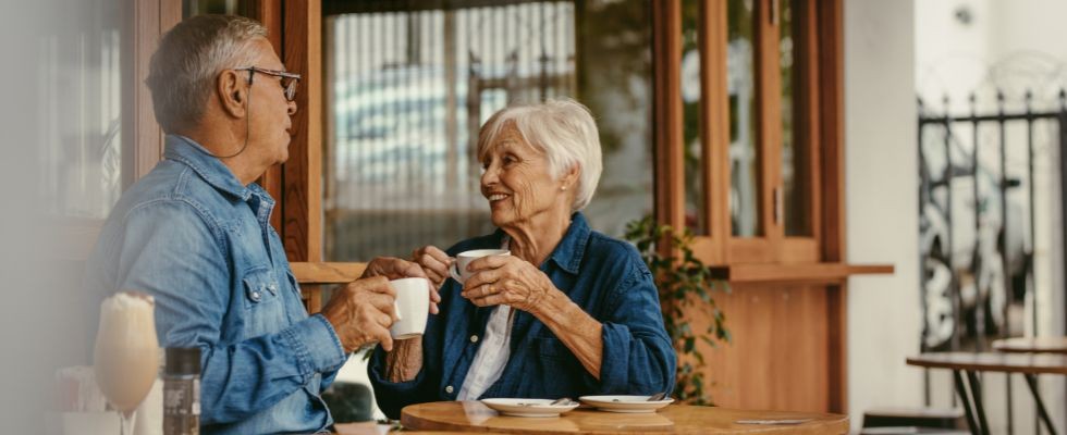 An elderly couple sits at the table drinking coffee and smiling