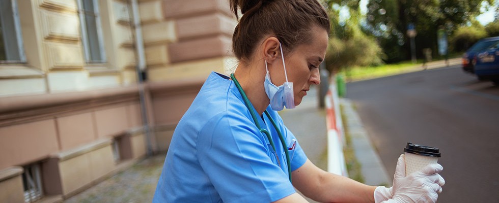 white female nurse looking sad. She has a blue mask under her chin and is holding a cup of coffee