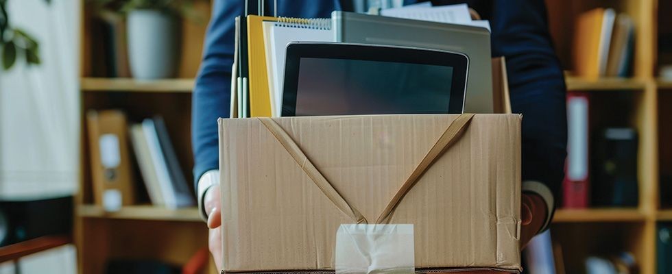 A man in a blue suit holds a brown box filled with office supplies.