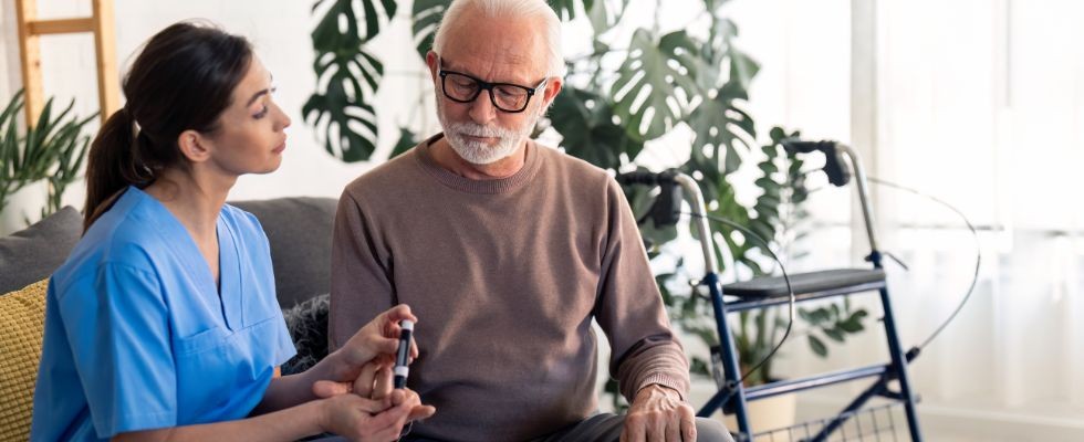 A Senior man looking at his hand while nurse is poking his finger with needle pen to measure his blood sugar level and sitting next to him during visit at home