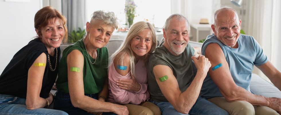 Five older individuals sit on a couch presenting their vaccine shot bandaids to the camera. They are all smiling.