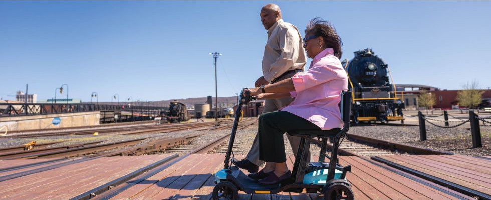 An elderly couple crossing train tracks. One is on a mobile scooter, the other is walking.