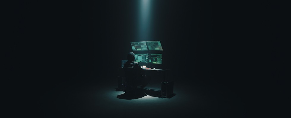 A person wearing a hoodie sits in front four computer monitors under a spotlight