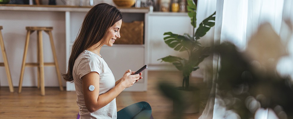A woman with a continuous glucose monitor on her arm sits on the floor looking at her phone while smiling