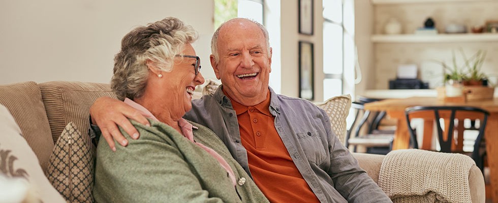 An elderly couple sit on the couch laughing