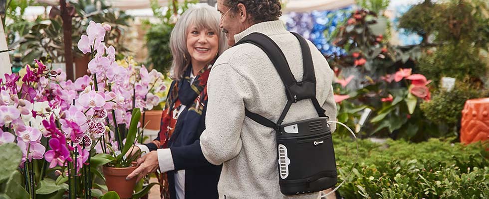 Two older people in a flower market, one is wearing a portable oxygen concentrator