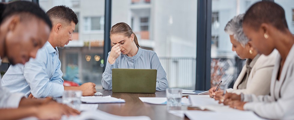 A woman sits at the head of a conference table pinching her nose with her laptop open