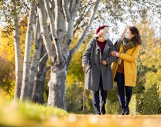 An elderly woman walks with a cane in one hand and her other arm linked around her caregiver's arm