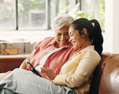 An elderly woman and her daughter sit on a couch smiling while looking at an iPad.
