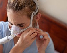 A woman puts on a CPAP machine and appears to be frustrated or in pain.