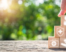 In the foreground a hand is stacking six blocks into a pyramid. On each block are different symbols of health care, including: a doctor, an ambulance, a syringe and pill, a medical shield, a first aid kid and a hand with a heart and pulse over it. In the background are blurry trees. 