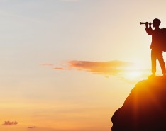 A man standing on a rock with a telescope looking out into sunset. 