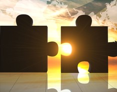 Foreground: Silhouettes of two people pushing together two large puzzle pieces. In the background is a sunset, shining through the gap in the puzzle pieces and a superimposed map of the world. 