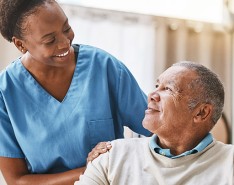 A caregiver smiles at a patient sitting down in a room.