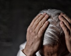 An elderly person with grey hair holds onto the top of their head.