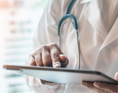 Image of person in white lab coat with stethoscope around their neck looking at and touching an ipad. 