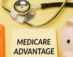 Orange basic calculator, yellow sticky note that says Medicare Advantage, piggy bank and stethoscope on pale yellow background