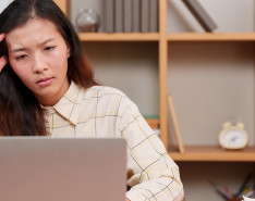 A young woman sitting at a desk staring at her laptop with her fingers to her temple.