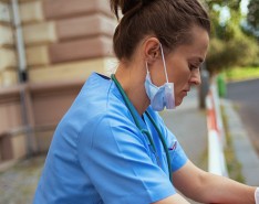 white female nurse looking sad. She has a blue mask under her chin and is holding a cup of coffee