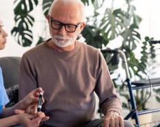 A Senior man looking at his hand while nurse is poking his finger with needle pen to measure his blood sugar level and sitting next to him during visit at home