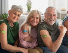 Five older individuals sit on a couch presenting their vaccine shot bandaids to the camera. They are all smiling.