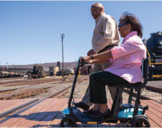 An elderly couple crossing train tracks. One is on a mobile scooter, the other is walking.