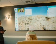 A room full of people watching a virtual reality simulation on a television monitor.