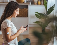 A woman with a continuous glucose monitor on her arm sits on the floor looking at her phone while smiling