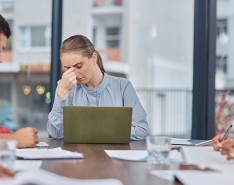 A woman sits at the head of a conference table pinching her nose with her laptop open