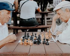 Two elderly men in white shirts playing a game of chess outside on a wooden table.