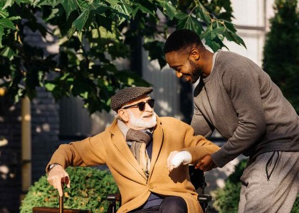 An elderly man in a wheelchair is helped by his caregiver